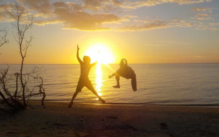 Two people on a beach leap into the air as the sun sets over the horizon across the water. One person appears to be mid-flip.